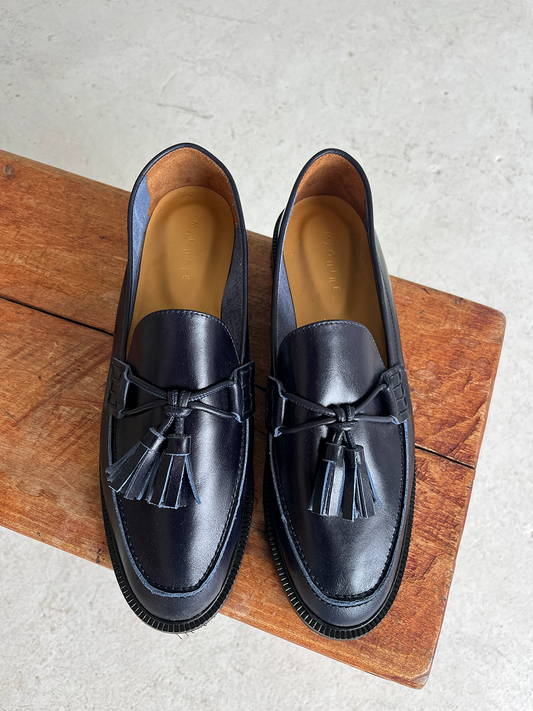 Pair of navy loafers with pompoms on wooden stool, white floor background