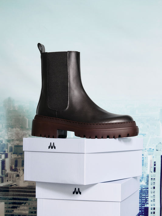 Dark brown Chelsea boot on a white box with a blurred cityscape background