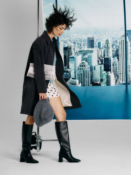 Person wearing a black coat, white shirt, polka dot skirt, and black knee-high boots with a cityscape in the background.