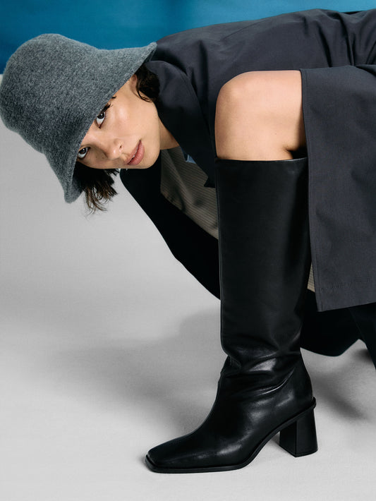 Person wearing a gray beanie, black coat, and black knee-high boots on a gray background