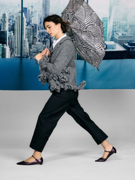 Woman in a gray blazer with ruffled sleeves and black pants walking against a cityscape background.