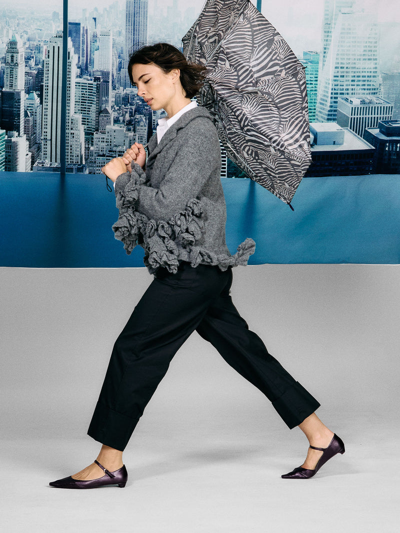 Woman in a gray blazer with ruffled sleeves and black pants walking against a cityscape background.