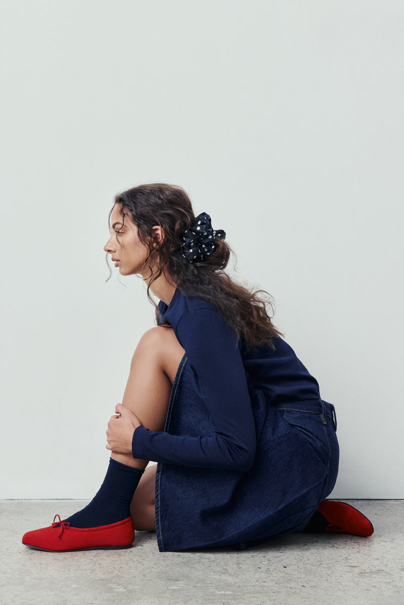 Woman sitting on the floor wearing a navy outfit, red shoes, and black socks against a plain background