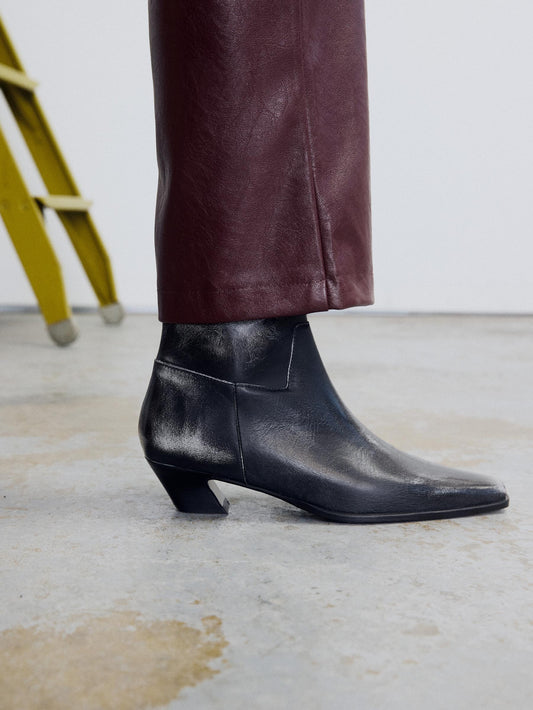 One foot of a two tone black ankle boot with a pointed square-toe and sculptural heel with a bordeaux pant