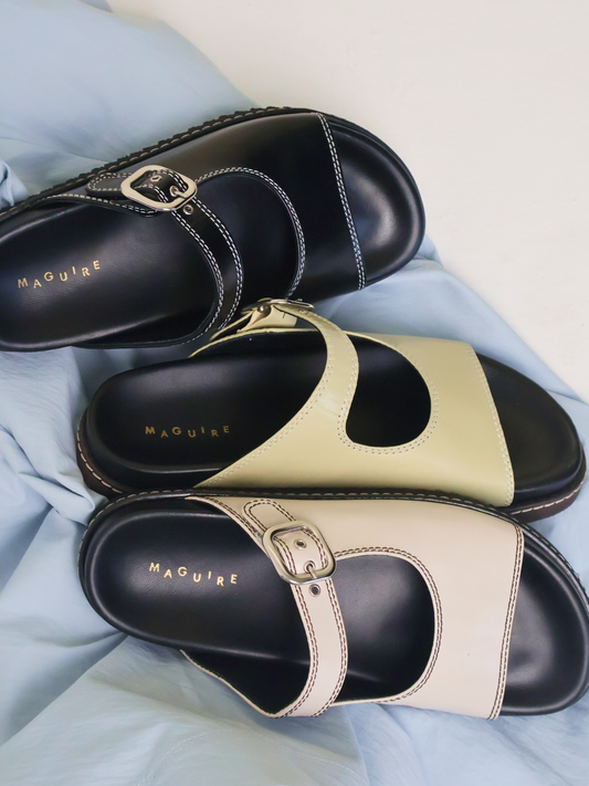 Pair of black and beige sandals with 'Maguire' branding on a light blue fabric background.