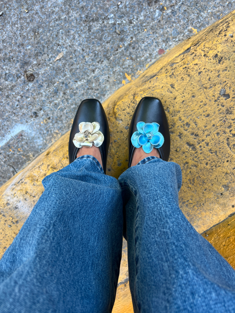Gold and blue metallic flower shaped shoe clips on black ballet flats worn by woman wearing jeans on a yellow step