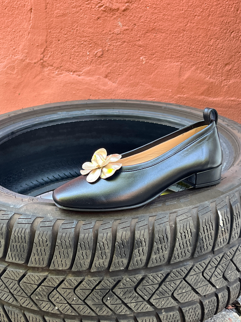 Gold metallic flower shoe clip on black ballet flat, standing on tire on a terracotta coloured background 