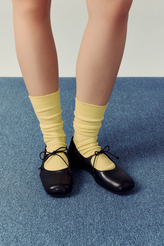 woman standing on blue carpet against white wall, wearing butter yellow socks and black mary jane ballet flats with lace tie