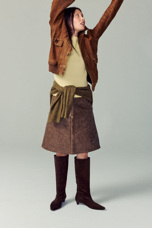 girl standing in white studio wearing brown suede heeled boots, brown skirt, yellow top with green sweater at her waist, smiling and playing around with jacket