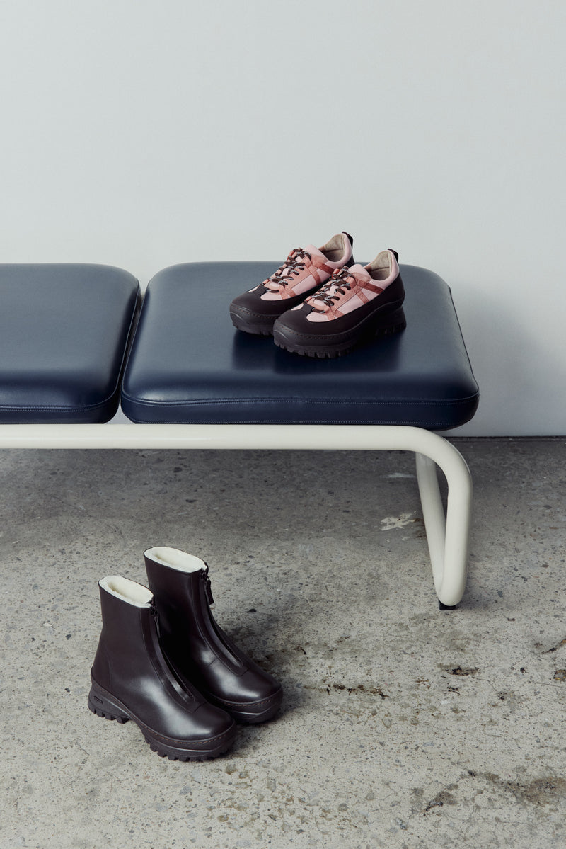 Pair of dk brown winter boots on a concrete floor with a bench in the background. The pink sneaker are on the bench.