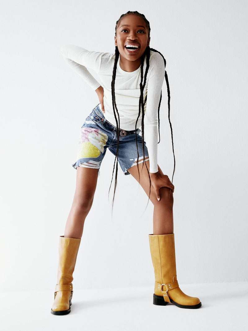 A yellow tan leather boot with a harness detail and low heel worn by a model wearing short.