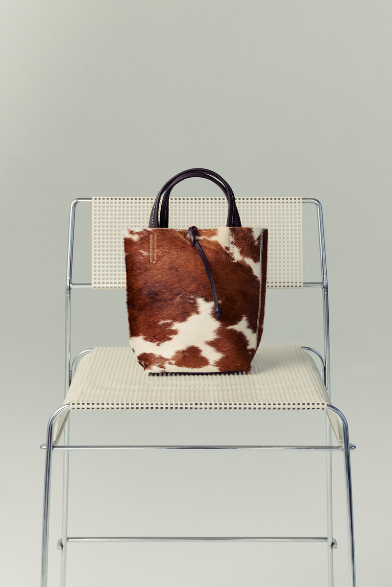 Brown and white cowhide Carpi leather bag on a clear chair with a gray background