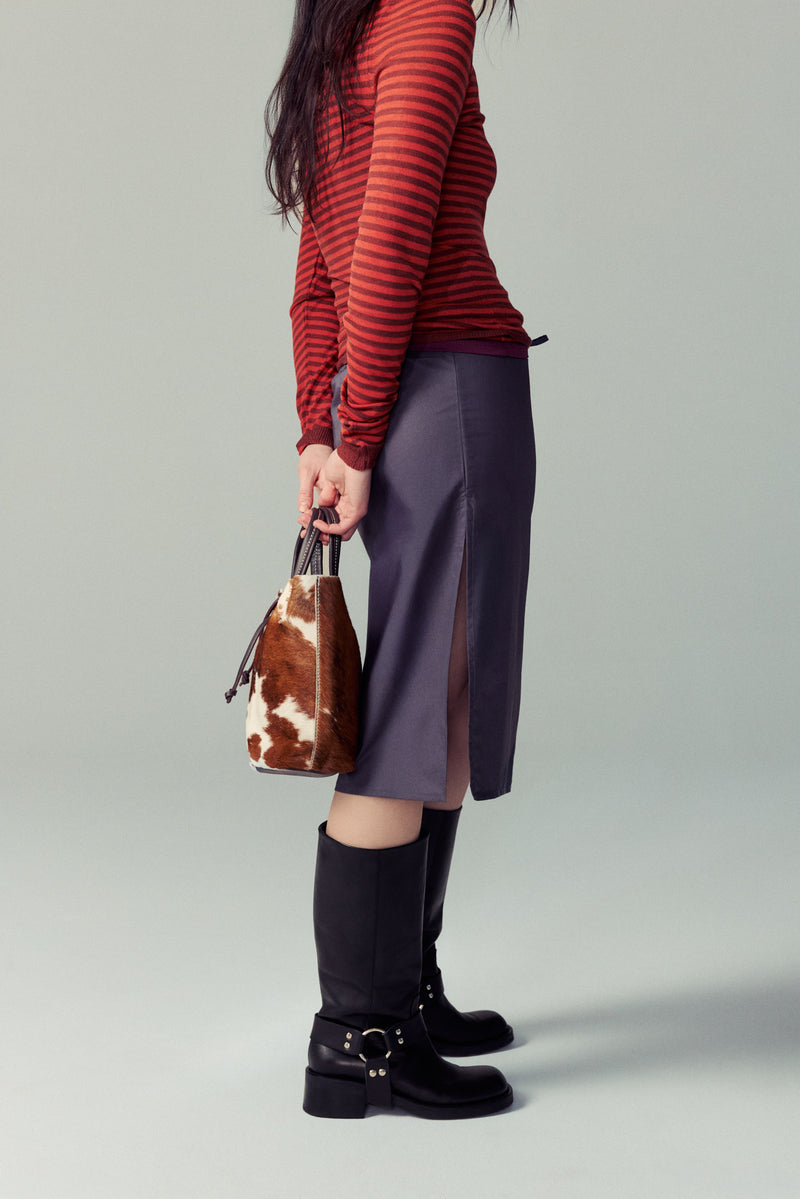Person wearing a red striped sweater, purple skirt, and black Lucca boots holding a cowhide handbag on a gray background