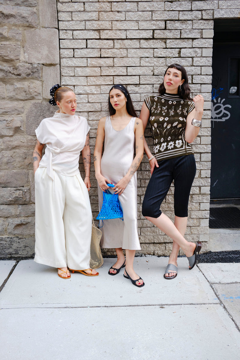 Three women standing against a stone wall, wearing stylish outfits.