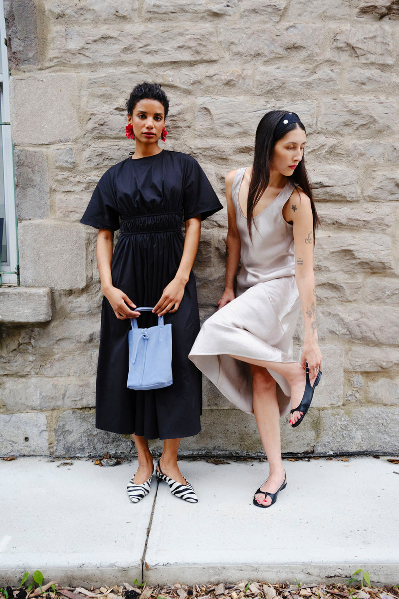Two women standing against a stone wall, one in a black dress with zebra shoes and the other in a beige dress with black sandals.