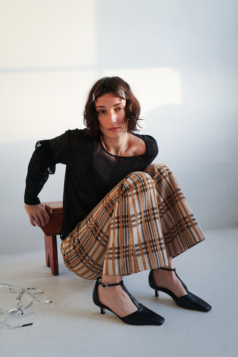 Woman leaning on a stool wearing a black top, plaid skirt and black t-strap kitten heels with a white background