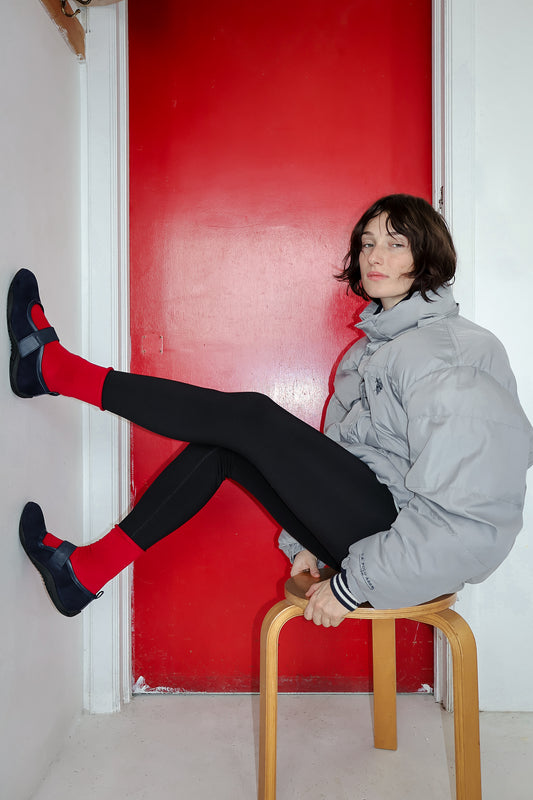girl sitting on stool against red door, wearing gray puffer coat, black leggings, red socks and navy flats.