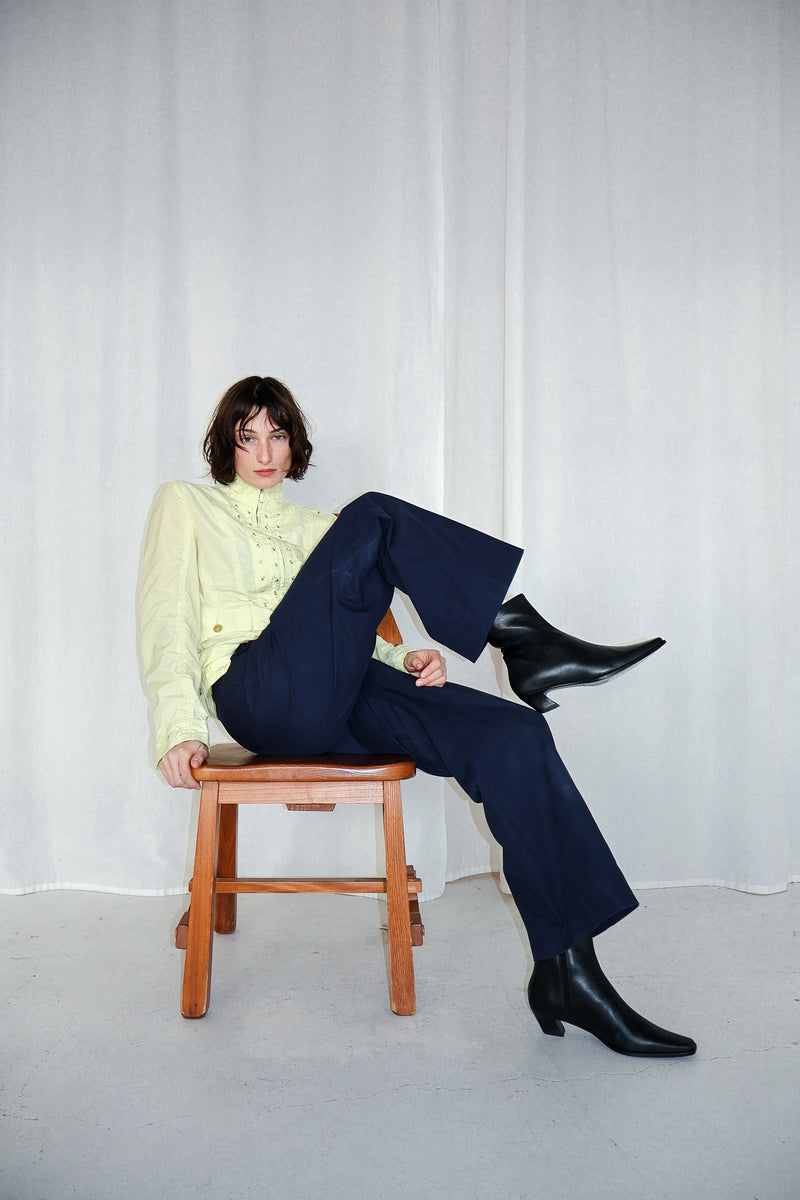 Person sitting on a wooden stool wearing a light yellow blouse, dark blue pants, and black boots against a white curtain background.