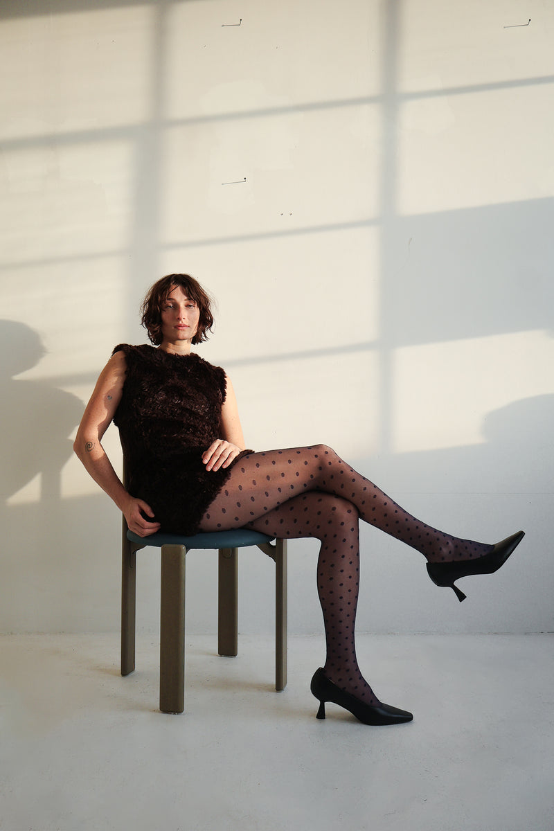 Woman sitting on a chair wearing a black dress and patterned stockings in a softly lit room.