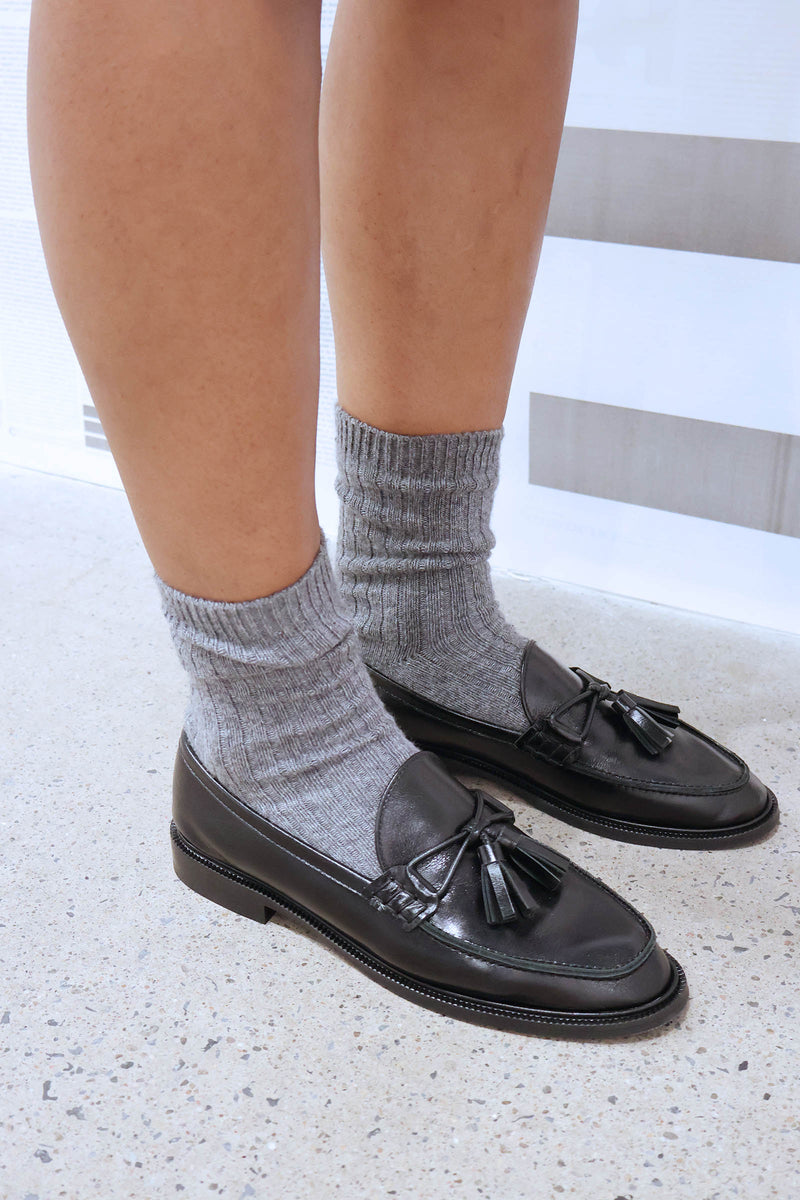 Black loafers with tassels worn with gray socks on a light background