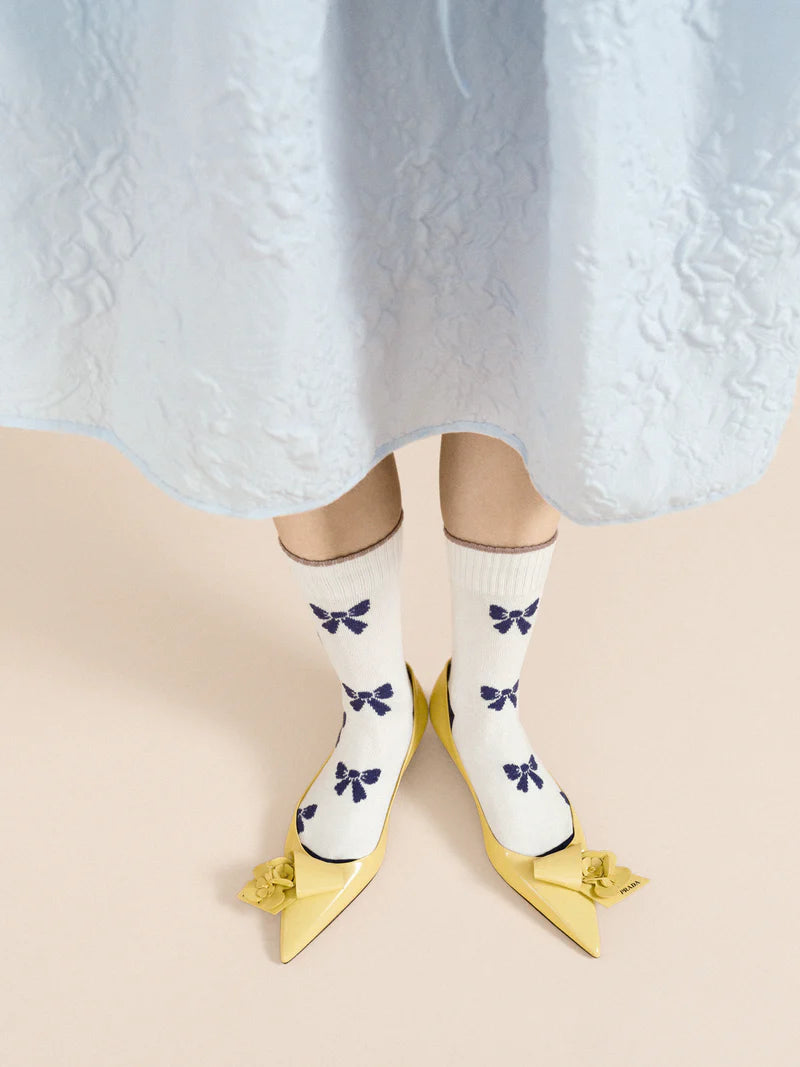 A pair of ivory crew socks with blue bows and yellow shoes, worn on a mannequin with a lacey blue dress .