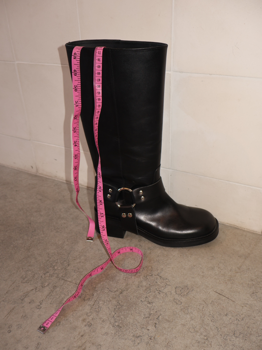A black leather boot with a low heel and a metallic ring detail on the shaft, designed for wider calves. There's a measuring tape on top.