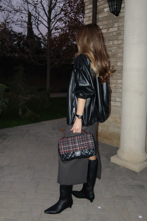 Woman in black leather jacket and boots holding a plaid handbag at night.