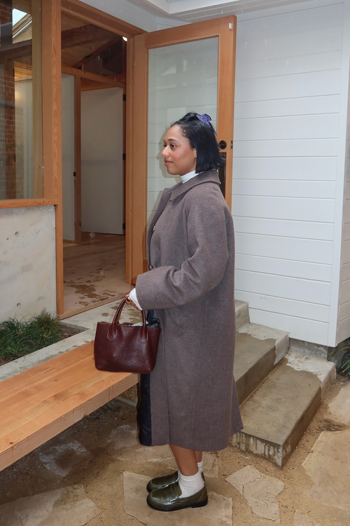 Woman in a long coat standing in front of a wooden door with a bench to her left with Orio Olive Loafers.