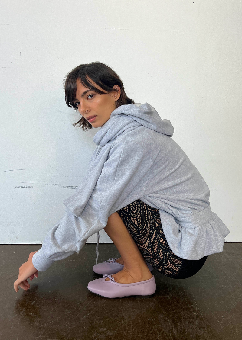Woman wearing a gray hoodie and black pants with lace details, sitting on a dark surface against a white wall.