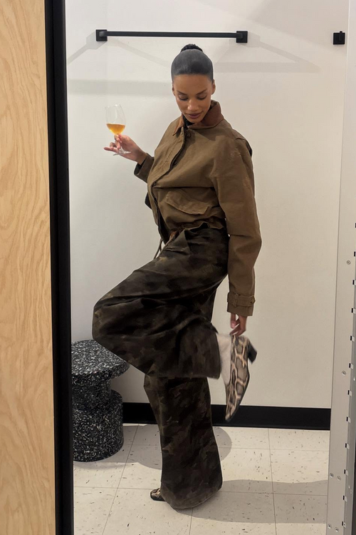 Woman in a brown outfit holding a drink in a room with a mirror with Soria Snake Heels.